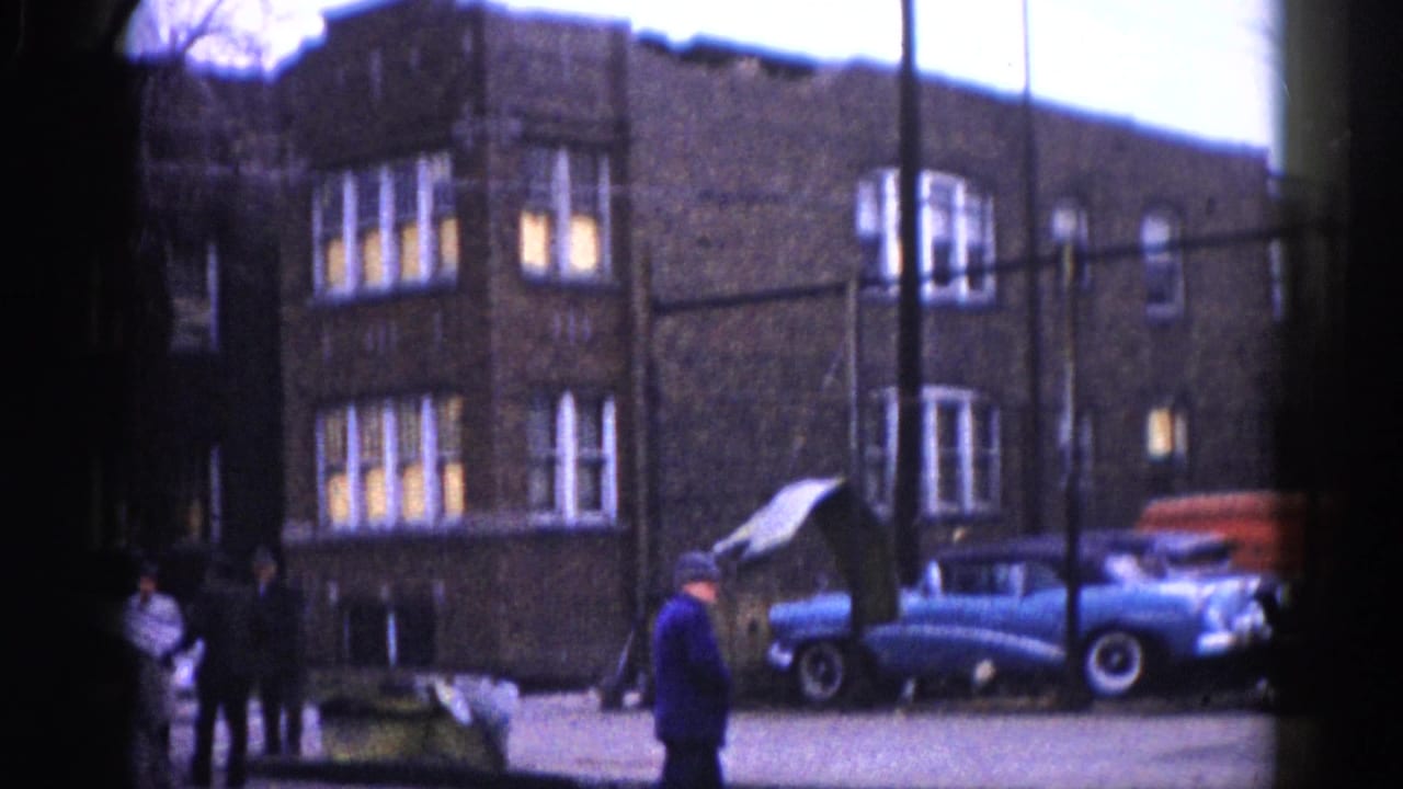 8mm home-movie frame circa 1962, Illinois: dark brick building with lit windows, a blue late-1950s American sedan at the curb, and pedestrians in winter coats.