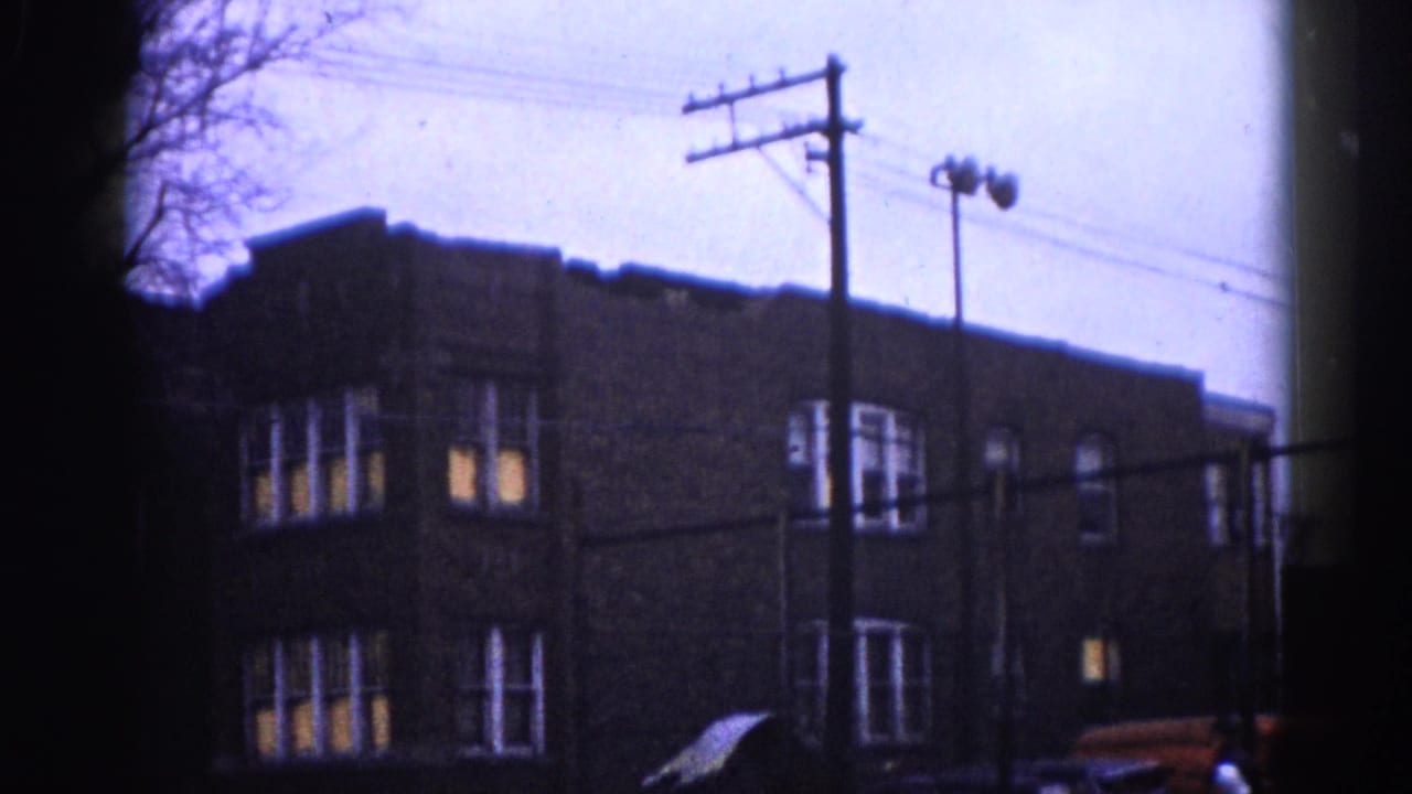 8mm home-movie frame circa 1962, Illinois: three-story brick building with lit windows and utility poles; a partial red vehicle visible at the lower right edge.