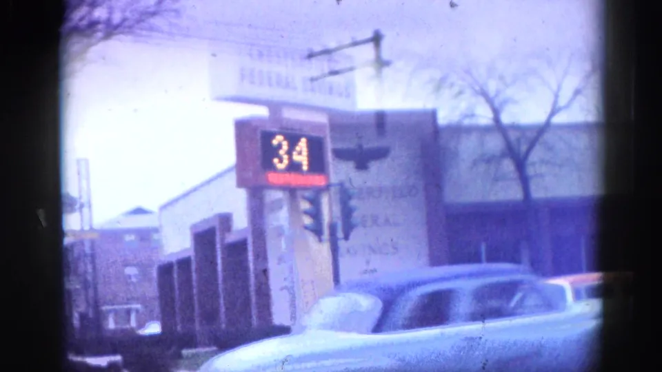 8mm home-movie frame circa 1962, Illinois: Chesterfield Federal Savings exterior with an early electronic temperature sign reading 34°F; a blue classic sedan in the foreground.
