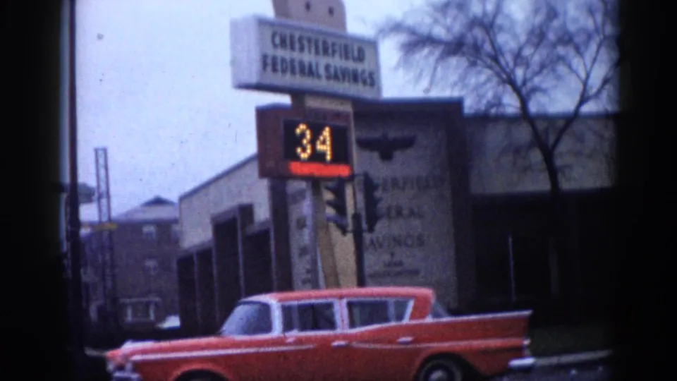 8mm home-movie frame circa 1962, Illinois: Chesterfield Federal Savings with an early digital temperature sign at 34°F; a red early-1960s station wagon and blue sedan at the curb.