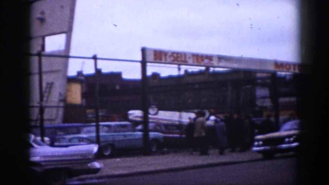 Street view of early 1960s American cars parked and driving, with a BUY-SELL-TRADE MOTORS sign visible in the background.
