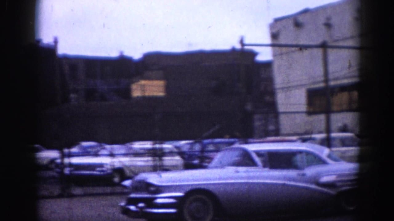 Illinois street, circa 1962, dominated by large chrome-trim American cars, with a partial BUY-SELL-TRADE MOTORS sign in the background.
