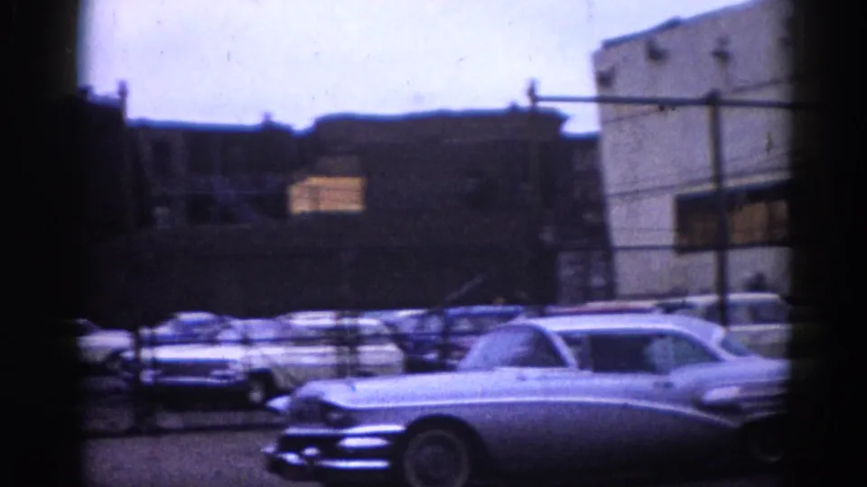 Illinois street, circa 1962, dominated by large chrome-trim American cars, with a partial BUY-SELL-TRADE MOTORS sign in the background.