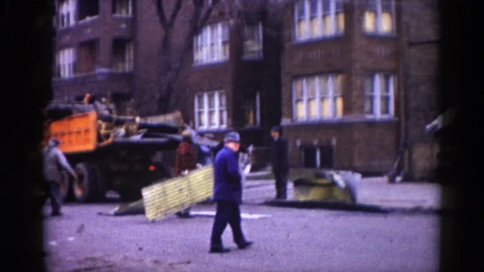 Illinois street construction scene, circa 1962: a worker crosses the foreground, an orange dump truck is parked left, and a brick apartment building with lit windows stands behind.