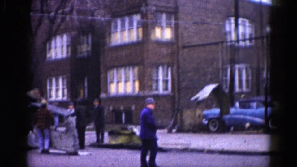 Illinois construction street scene, circa 1962: workers, construction debris, a brick apartment building with lit windows, and a small blue car parked at right.