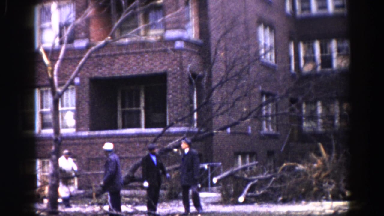 Men in dark winter coats gathered on a sidewalk outside a multi-story brick building, bare trees visible.