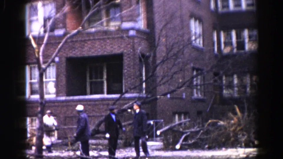 Several men in dark winter overcoats stand on a sidewalk in front of a multi-story brick building; bare trees suggest late autumn or winter, Illinois, circa 1962.