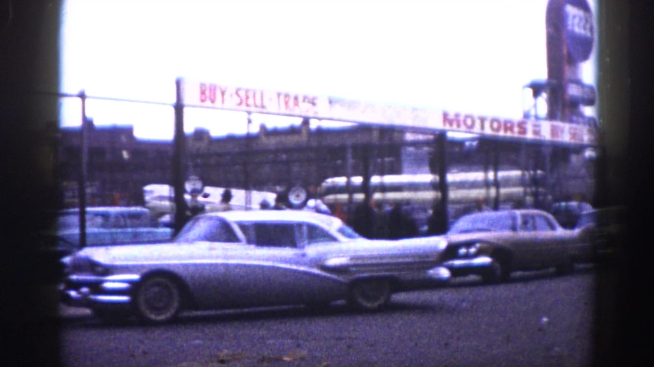 Silver tailfin American sedan at a used car lot with a "BUY SELL TRADE MOTORS" sign visible in the background.