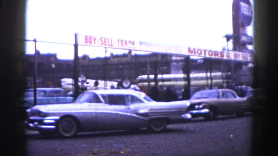 A large silver tailfin American sedan dominates the foreground of a used car lot; a "BUY SELL TRADE MOTORS" banner sign and a tall background sign are visible, Illinois, circa 1962.