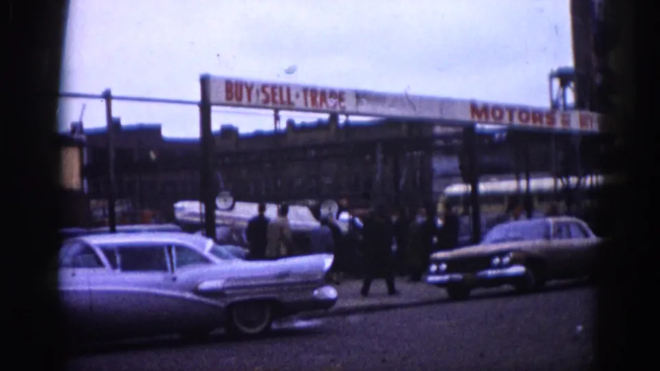 Groups of men in dark overcoats browse classic American tailfin cars at an open used car lot; a "BUY SELL TRADE MOTORS" sign spans the upper frame, Illinois, circa 1962.