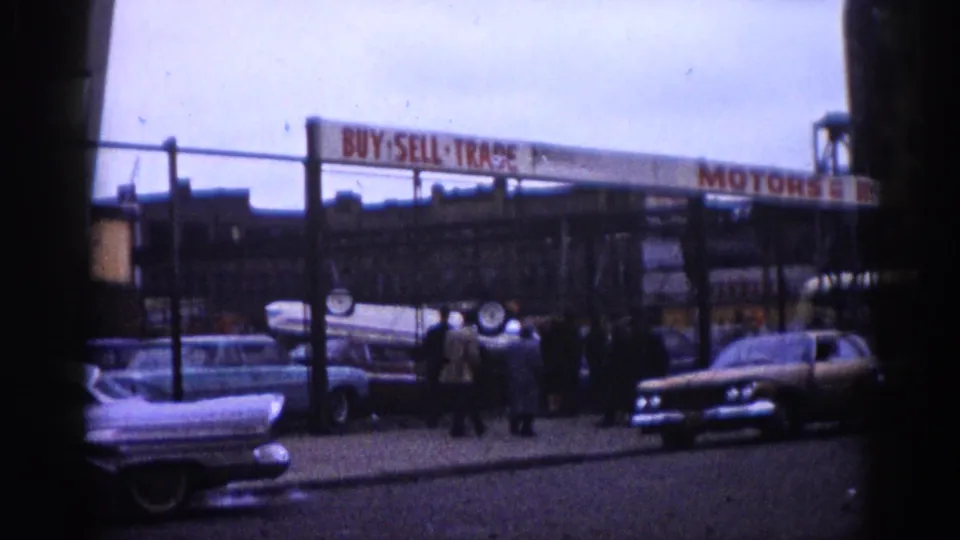 Wide view of a used car lot showing multiple finned American sedans; "BUY SELL TRADE MOTORS" sign is prominent, with groups of people browsing between vehicles, Illinois, circa 1962.