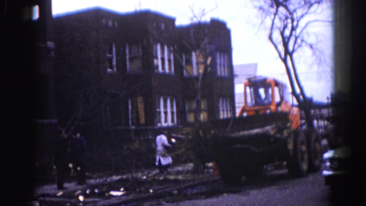 Orange front-end loader clears fallen tree branches on a residential street; multi-story brick buildings and workers visible in background.