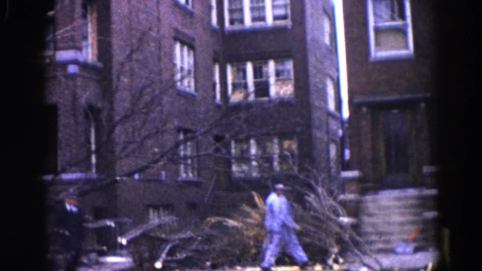 Workers move through storm debris and fallen branches in front of brick apartment buildings, residential Illinois neighborhood, circa 1962.