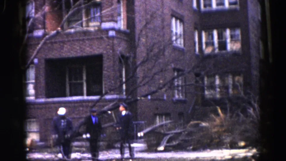 Group of three people amid storm debris in front of a brick residential building, Illinois, circa 1962; one wears a hat, another a lighter jacket.