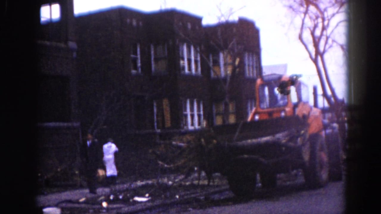 Orange front-end loader moves downed trees in front of a multi-story brick building; two figures stand amid street debris.