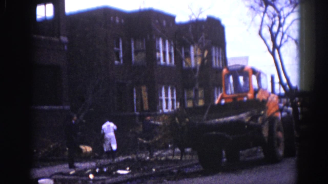 8mm frame circa 1962, Illinois — a figure in white coat and another in dark clothing survey downed trees as an orange front-end loader operates beside a multi-story brick building.
