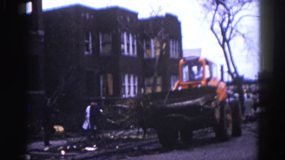 8mm frame circa 1962, Illinois — orange front-end loader moves a large fallen tree trunk while a white-coated figure works among scattered branches; multi-story brick building in background.