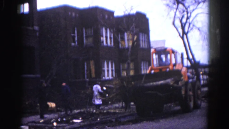 8mm frame circa 1962, Illinois — orange loader transports downed timber while a white-coated figure stands amid storm debris outside a multi-story dark brick building.
