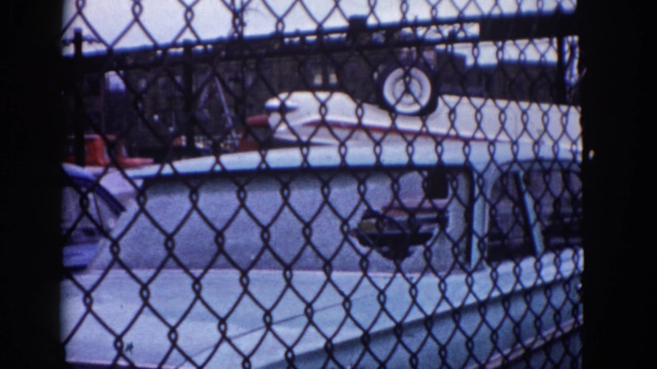 Chain-link fence obscures a light-colored automobile with red reflectors or taillights visible at frame edge; Illinois urban clearance scene on 8mm, circa 1962.