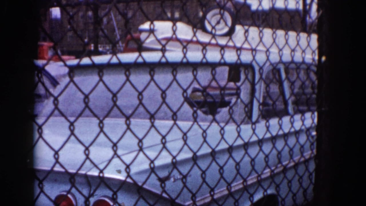 Fence-and-vehicle scene continues on 8mm; light-colored American car body visible through chain-link with red objects at frame edge, Illinois, circa 1962.