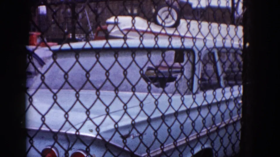 Fence-and-vehicle scene continues on 8mm; light-colored American car body visible through chain-link with red objects at frame edge, Illinois, circa 1962.