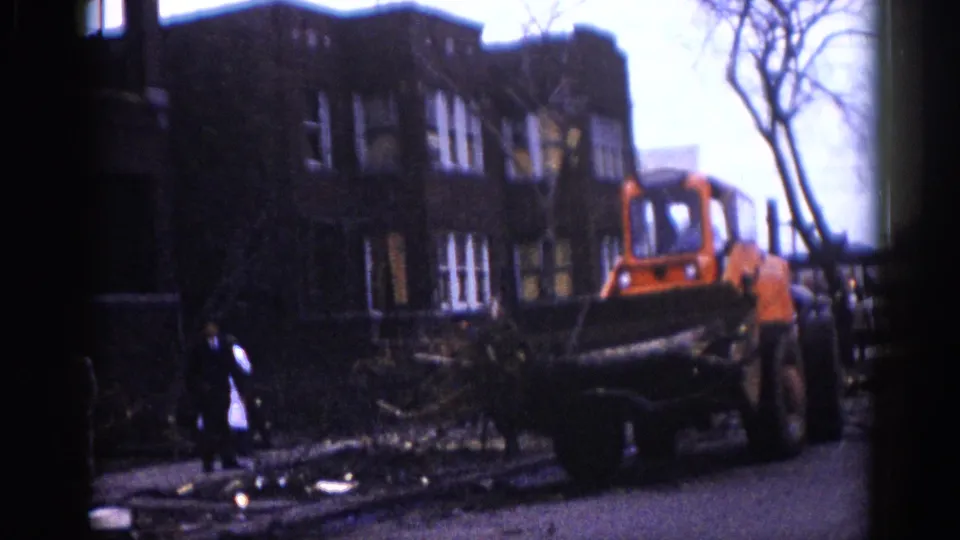 Orange front-end loader positioned beside a dark multi-story brick building with bare deciduous trees; a human figure stands nearby on debris-covered ground, Illinois, circa 1962.