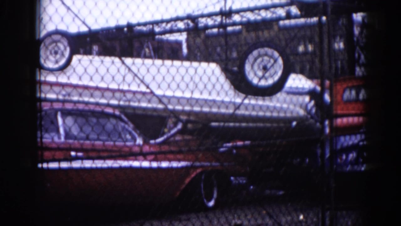 View through chain-link fence of a dark red sedan with a white car stacked inverted above it in a junkyard