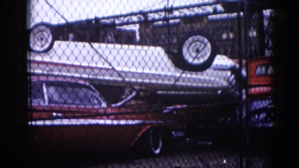 8mm film, circa 1962, Illinois: chain-link fence foreground framing a dark red American sedan and a white car stacked upside down with tires skyward in a salvage yard