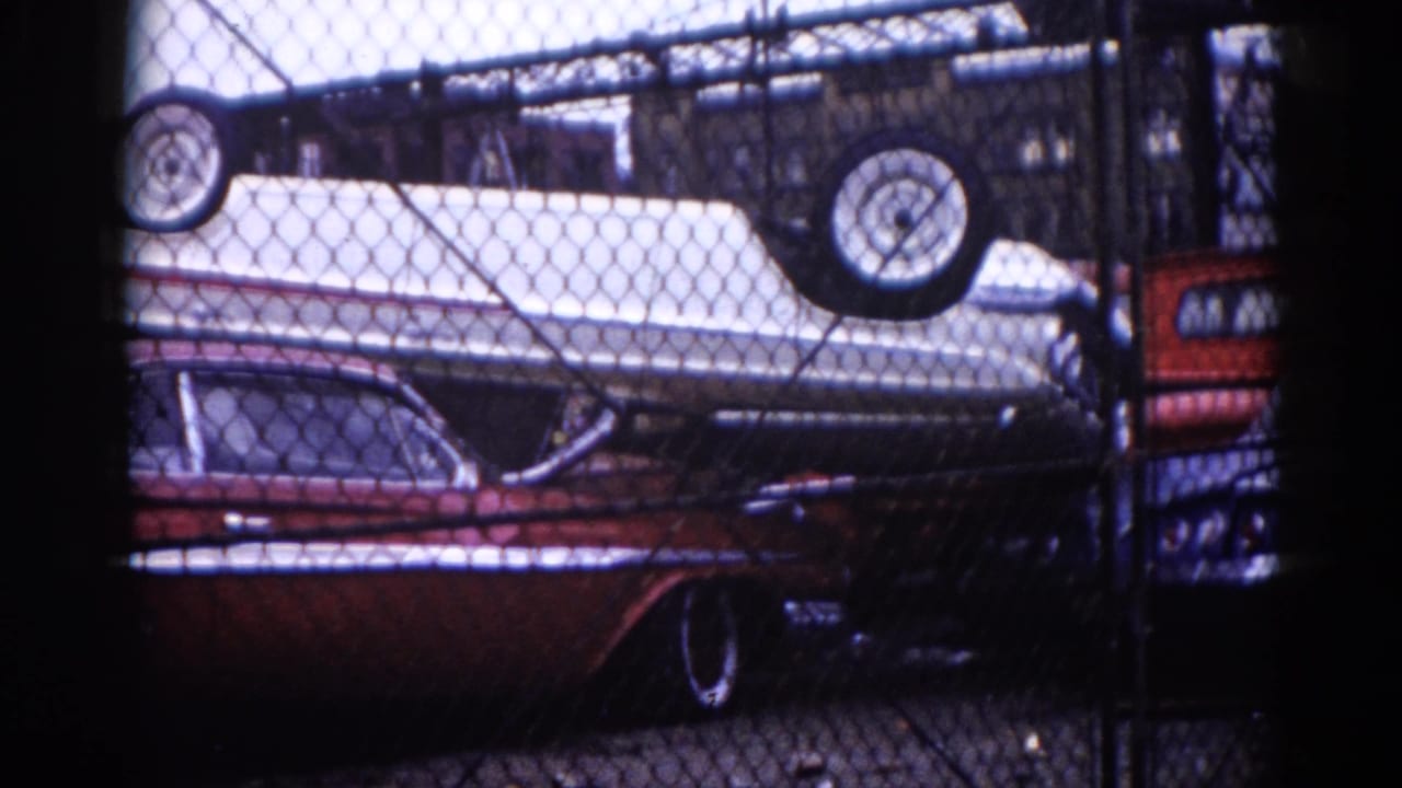 Chain-link fence view of dark red sedan with a white car stacked inverted above in salvage yard, slight camera shift