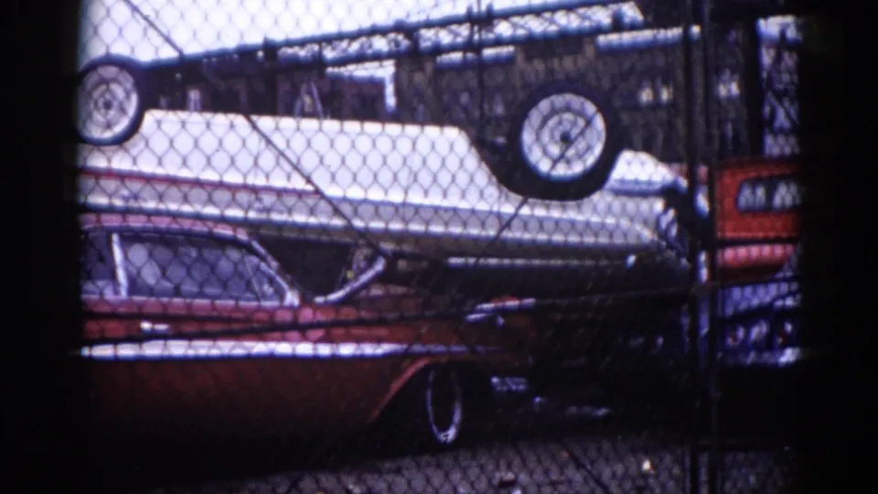 8mm film, circa 1962, Illinois: chain-link fence foreground with dark red American sedan and white car stacked inverted, tires skyward — minor camera movement from opening frame