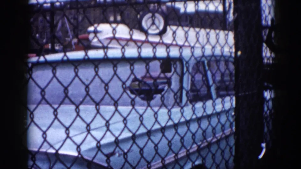 8mm film, circa 1962, Illinois: camera pans lower to reveal a pale blue car body through the fence below the stacked red and white vehicles