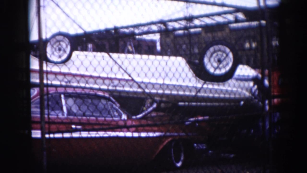 1962 Illinois 8mm footage: white boat hull upside down with wheels atop a dark red early-1960s American sedan, chain-link fence foreground, overexposed sky above.