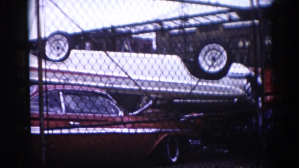 1962 Illinois 8mm footage: white boat hull upside down with wheels atop a dark red early-1960s American sedan, chain-link fence foreground, overexposed sky above.