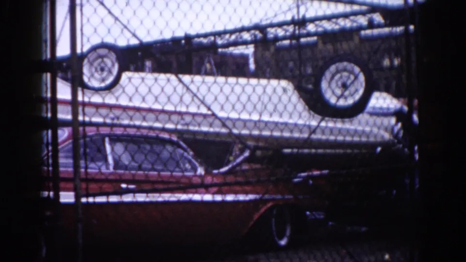 8mm home movie, Illinois c.1962: wider shot of inverted boat on red American sedan behind chain-link fence, rear of red car and industrial structures visible beyond.