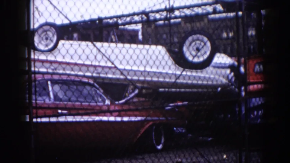 Final 8mm frame, Illinois 1962: inverted boat on dark red sedan behind chain-link fence, partial colored panel visible at right edge of frame.