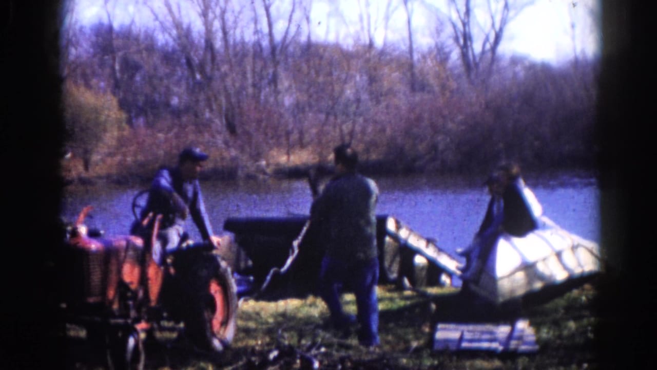Three men in 1960s work jackets handle equipment beside a red tractor on an Illinois riverbank in early spring or late fall.