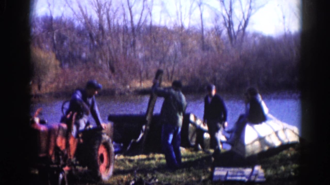 Work crew with a red tractor on a riverbank in what appears to be 1960s Illinois; men actively handle a large white hull or container.