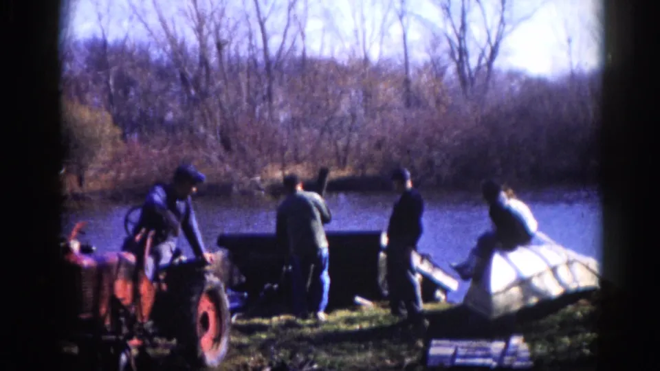Work crew near a red tractor; one man is seated atop a large white structure on an Illinois riverbank, circa 1962.