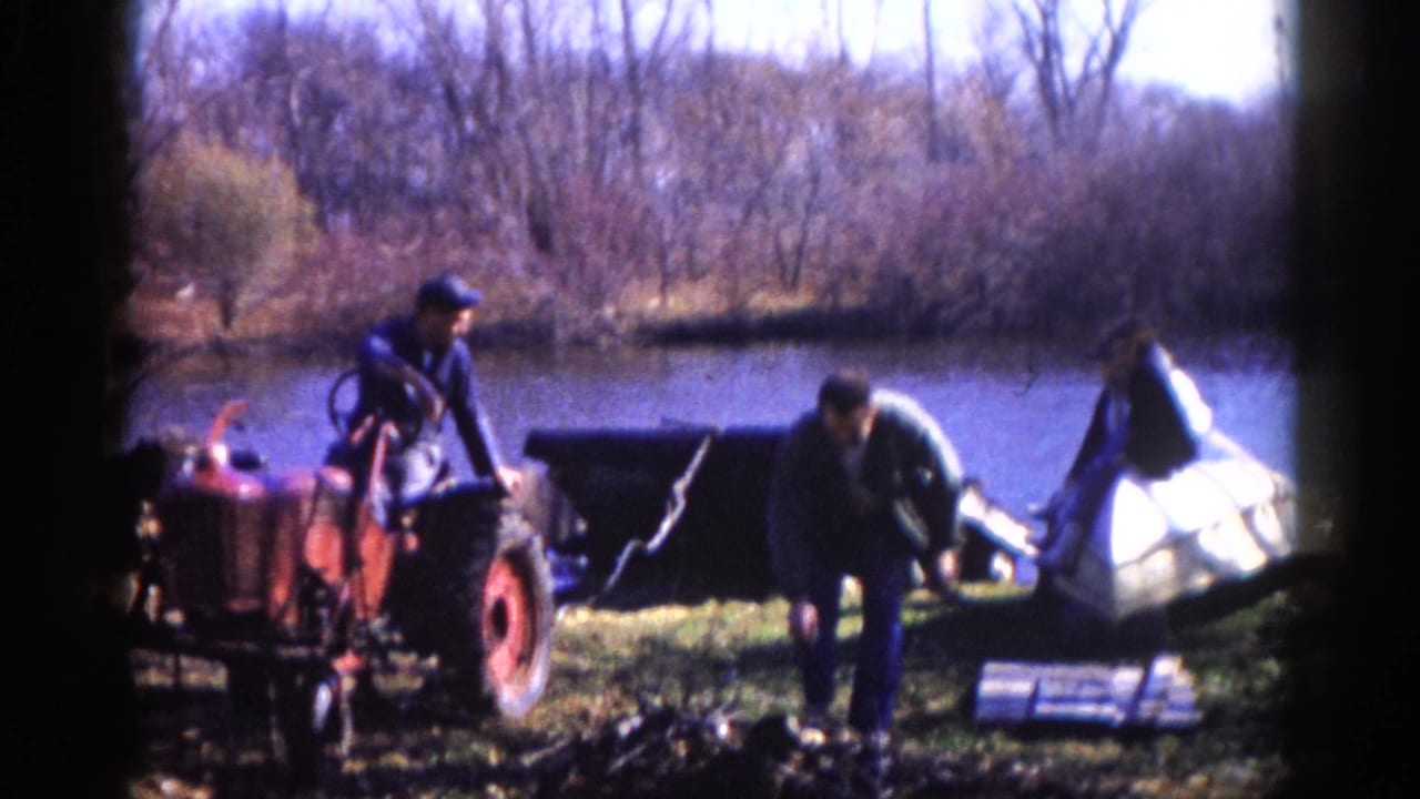Two men beside a red compact tractor at a pond bank in what appears to be rural Illinois, circa 1962, with a white boat hull and bare deciduous trees in the background.