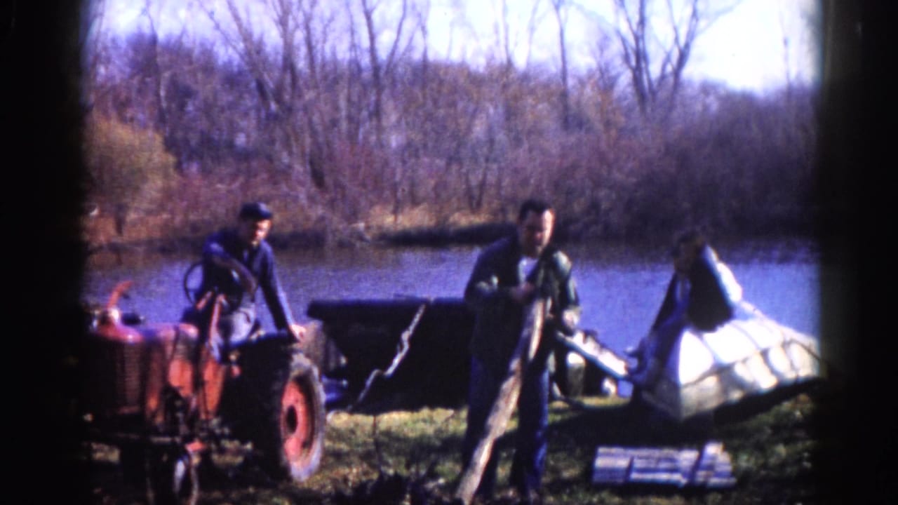 Two men positioned around a red compact tractor and a white boat hull on a rural pond bank, possibly Illinois circa 1962, with bare trees visible across the still water.