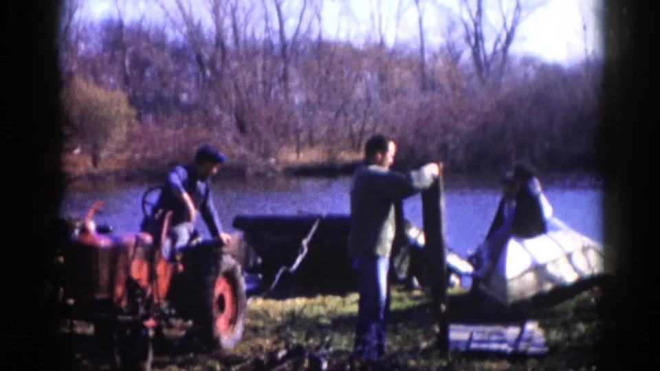 Two men in work clothing maneuvering a white boat hull beside a red tractor on a pond bank, rural Illinois area, circa 1962, bare trees visible in the background.