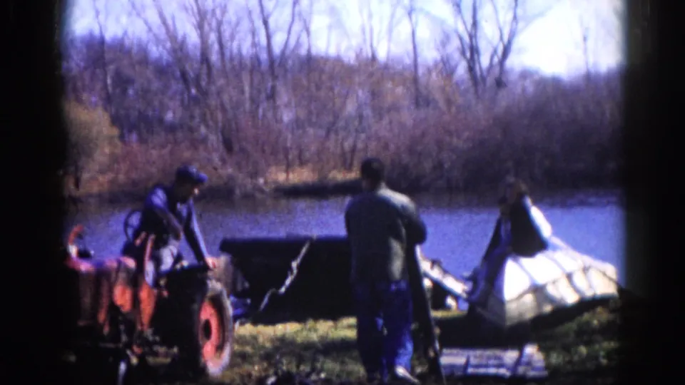 Two men continuing to work with a white boat hull and red tractor at a rural pond bank, consistent with rural Illinois home-movie footage circa 1962.