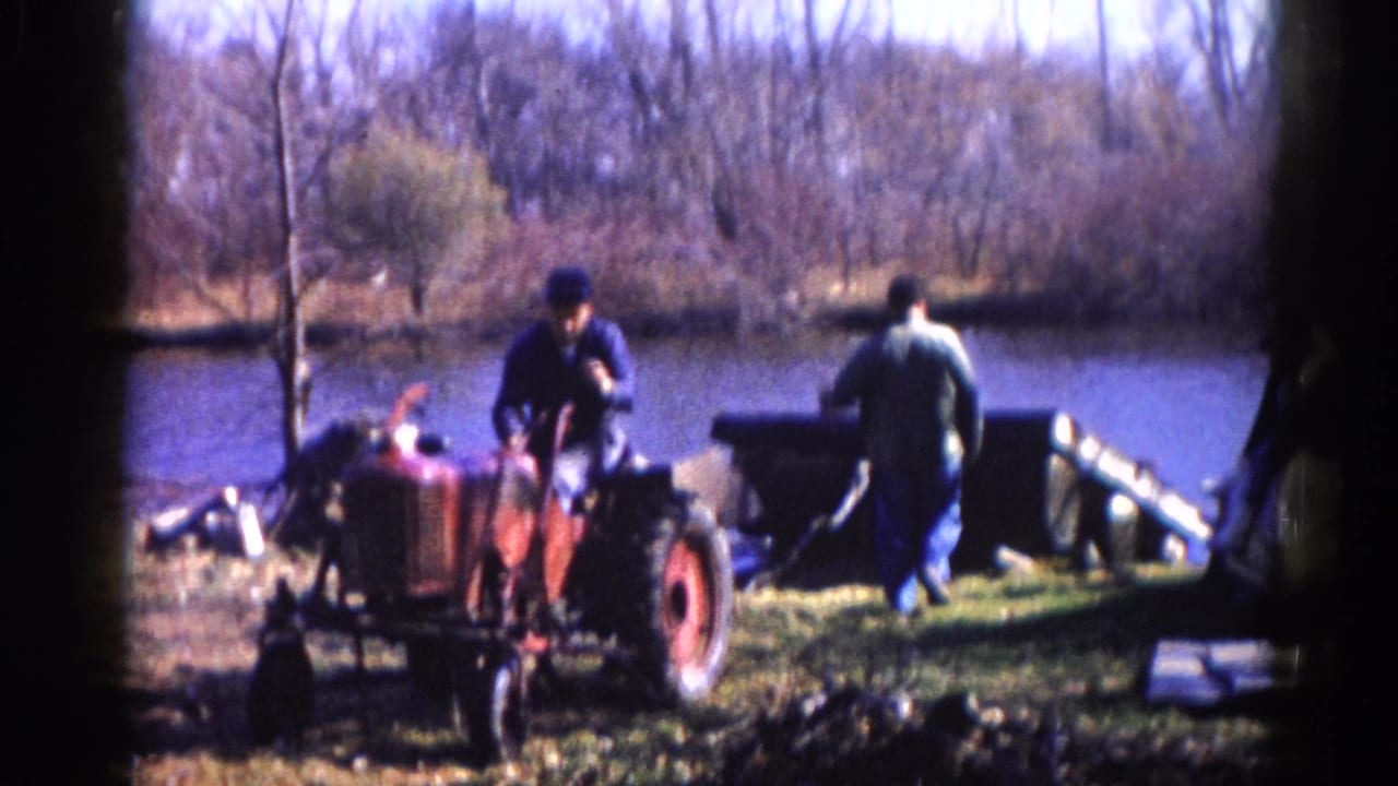 Two men working near a compact red tractor at a lake's edge, bare trees lining the far shore.