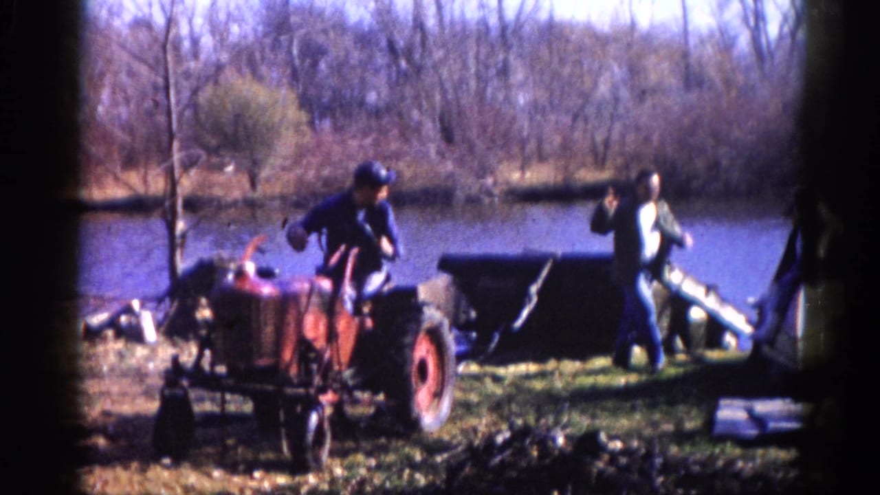 Two men in work clothing alongside a compact red tractor at a rural lakeshore, bare deciduous trees and calm water visible, rural Illinois circa 1962, 8mm film.