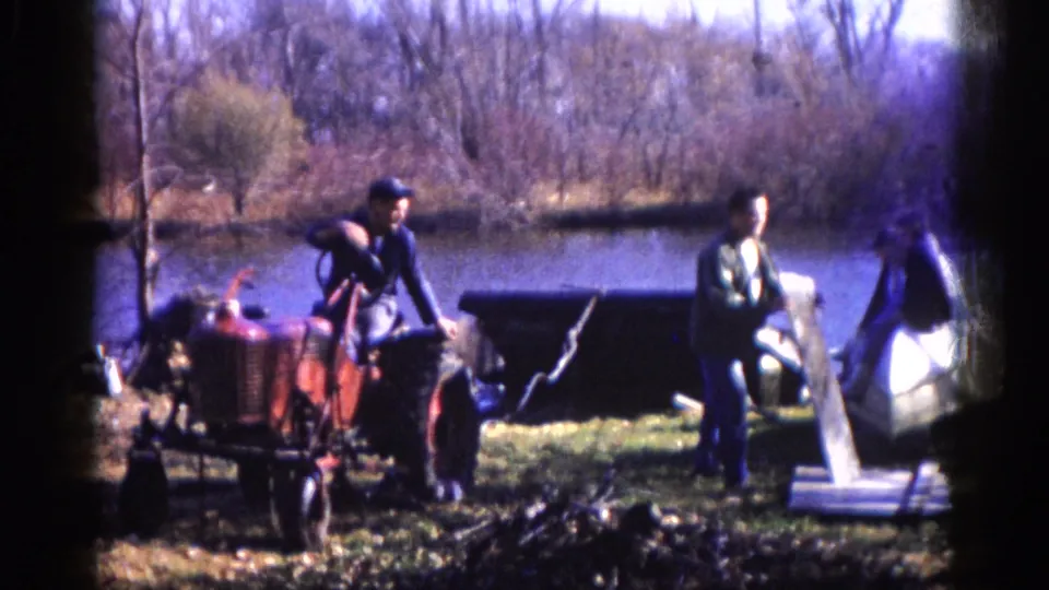 Closer view of two men and a compact red tractor at a rural lakeside bank; a pale tarp-like material appears at the water's edge, circa 1962 Illinois, 8mm film.