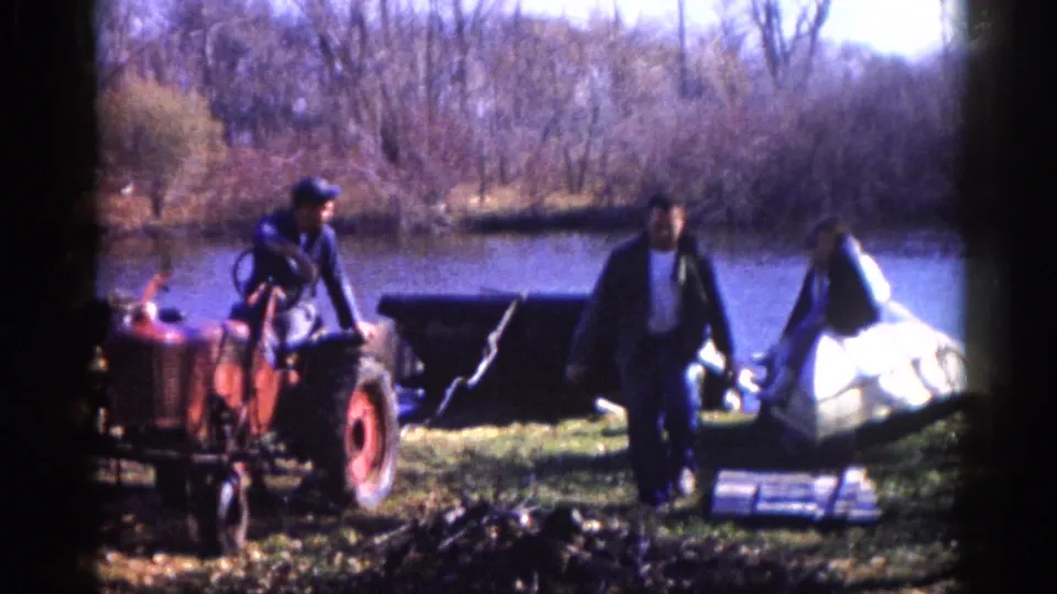 Final frame shows two men completing a retrieval task with a compact red tractor at a bare-treed lakeside in apparent late autumn, circa 1962, rural Illinois, shot on 8mm film.