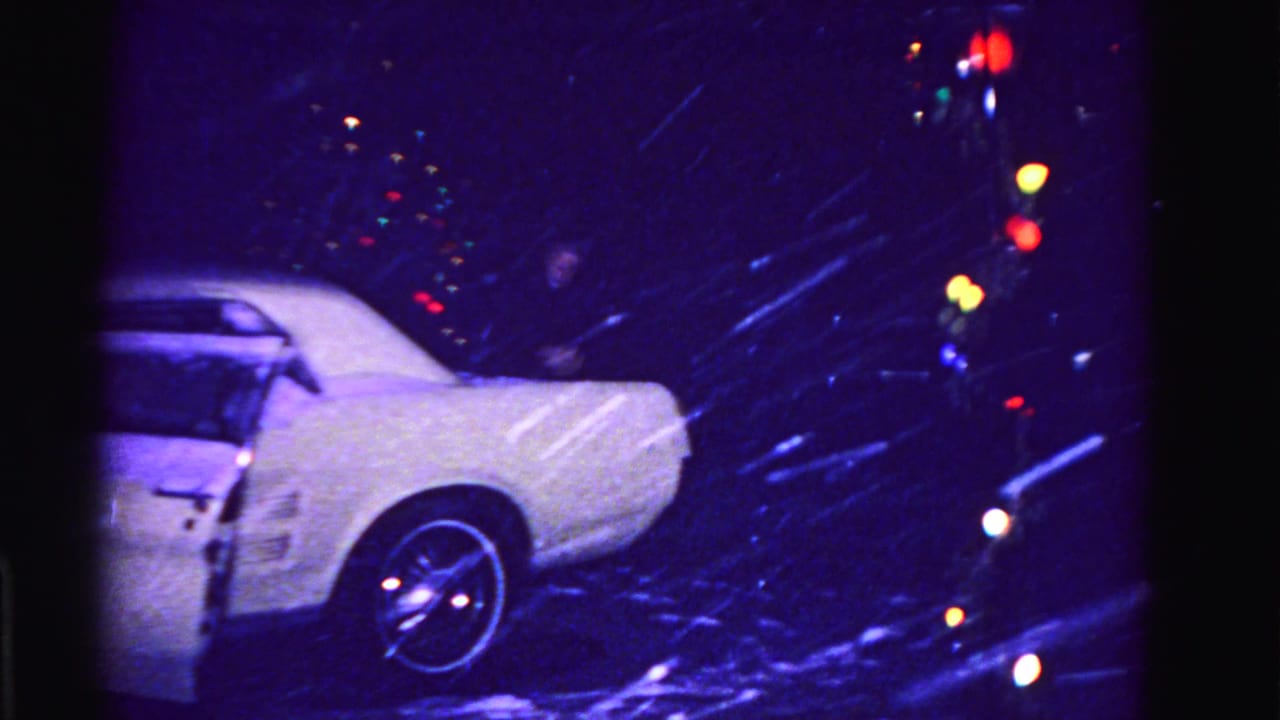 Snow-covered white American car in a residential driveway at night, circa 1962 Hagerstown Maryland, multicolored incandescent holiday lights glowing behind, 8mm film.