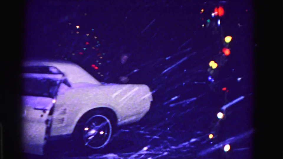 Snow-covered white American car in a residential driveway at night, circa 1962 Hagerstown Maryland, multicolored incandescent holiday lights glowing behind, 8mm film.