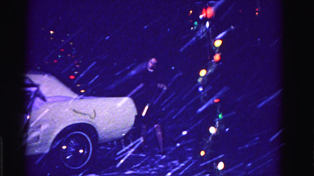 Woman in dark coat near the rear of a snow-covered white car at night, circa 1962 Hagerstown Maryland, multicolored Christmas lights glowing behind, 8mm archival footage.