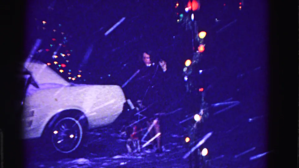 Woman in dark coat beside a snow-covered white car at night with a small dog in a red coat nearby, circa 1962 Hagerstown Maryland, multicolored holiday lights behind, 8mm film.